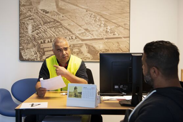 Twee mensen praten en zitten aan bureau.