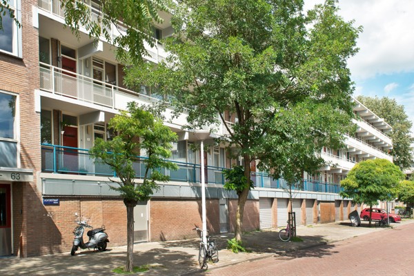 Straatbeeld met een meerlaagse flat met balkons, bomen langs het trottoir, geparkeerde fietsen, een scooter en een rode auto. Huisnummer 63 zichtbaar bij de ingang.