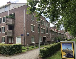 Appartementencomplex met veel groen ervoor en een trottoirbord met de tekst 'Koninklijk Paleis Amsterdam'.