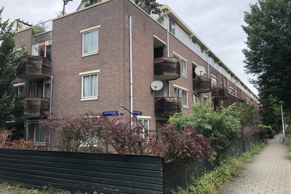 Hoekzicht op een appartementencomplex met rode bakstenen, houten balkonpanelen, schotelantennes en groen langs het trottoir.