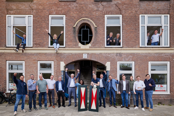 Groepsfoto voor een oud bakstenen gebouw, mensen staan buiten en zitten in open ramen boven, mogelijk tijdens een evenement.