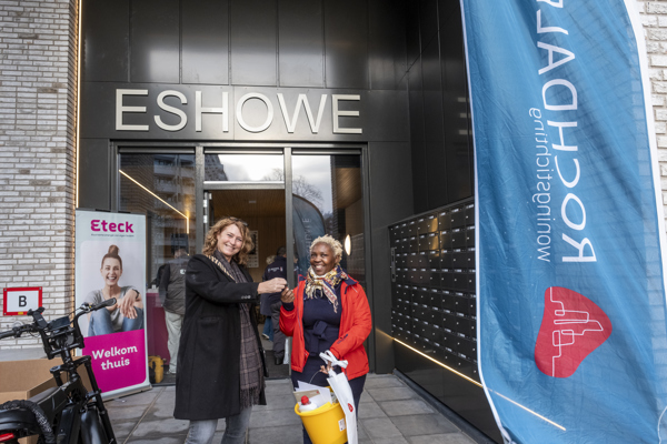 Voor de entree van een gebouw met de naam Eshowe overhandigt een medewerker sleutels aan een nieuwe bewoner. Voor de ingang staan mensen, een fiets en een grote vlag met het logo van Rochdale.